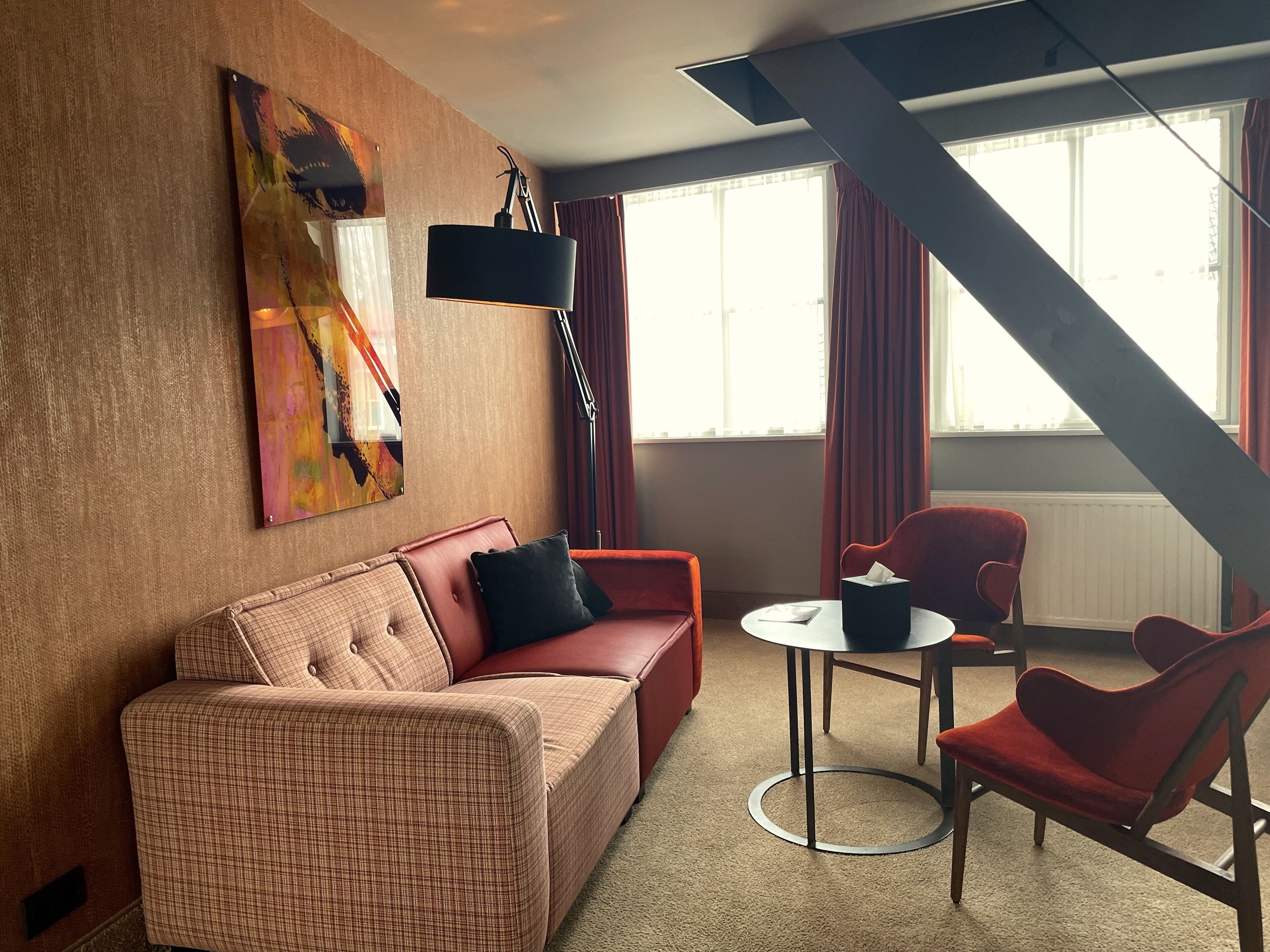 Une chambre d’hôtel moderne avec un canapé à carreaux, un fauteuil rouge, une petite table ronde et un grand tableau abstrait accroché au mur.