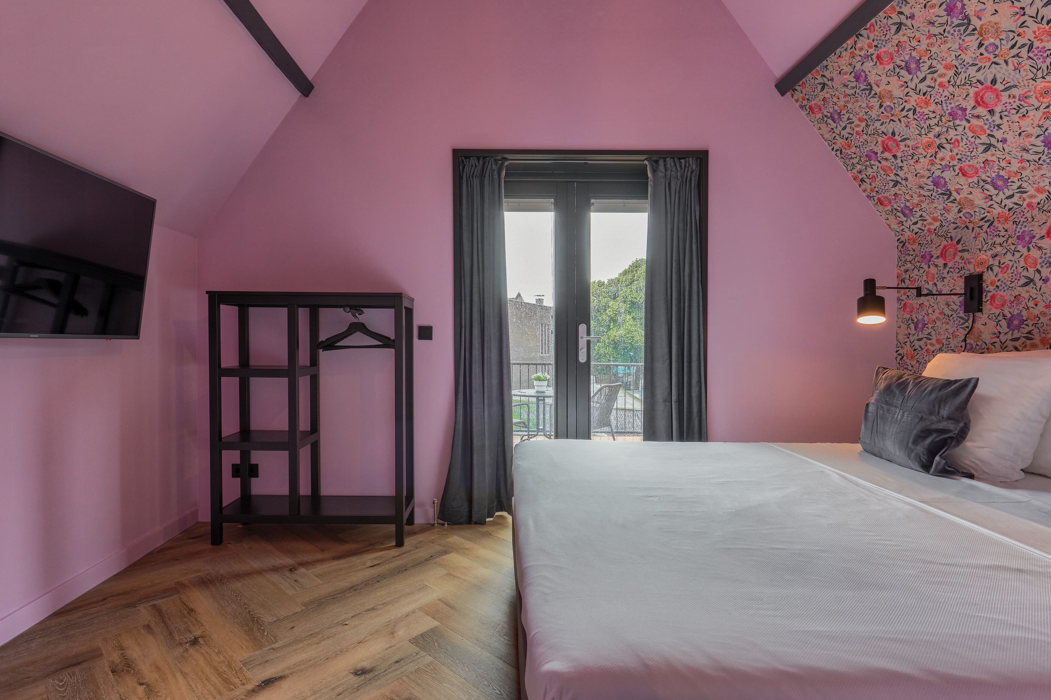 A bedroom with a bed, a wooden floor, a black hanger rack, a window with curtains, and a balcony with a table and chairs.