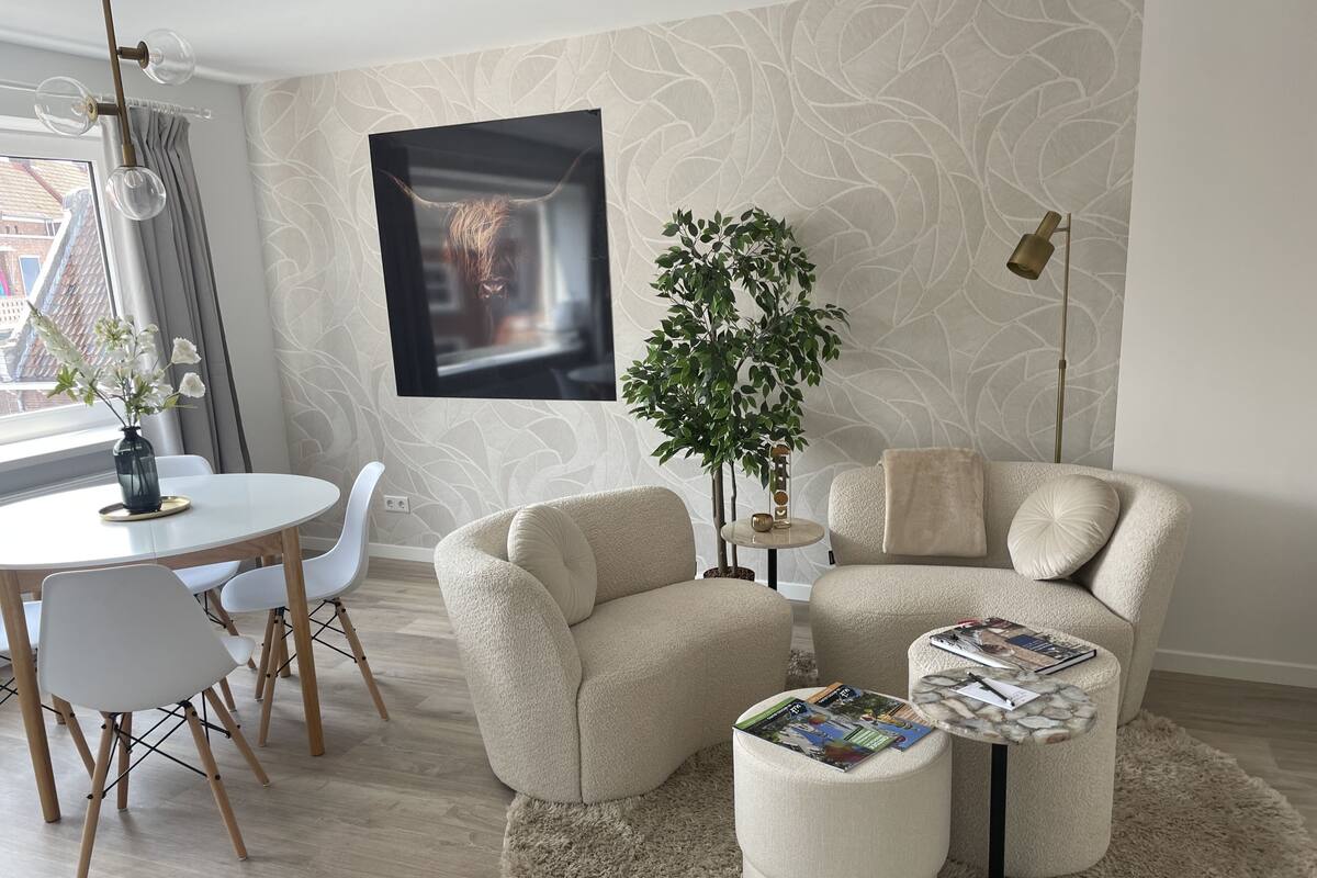 Un salon moderne avec une table de salle à manger ronde, deux fauteuils beiges, un petit pouf rond sur lequel sont posés des magazines, une plante en pot et un tableau accroché au mur.
