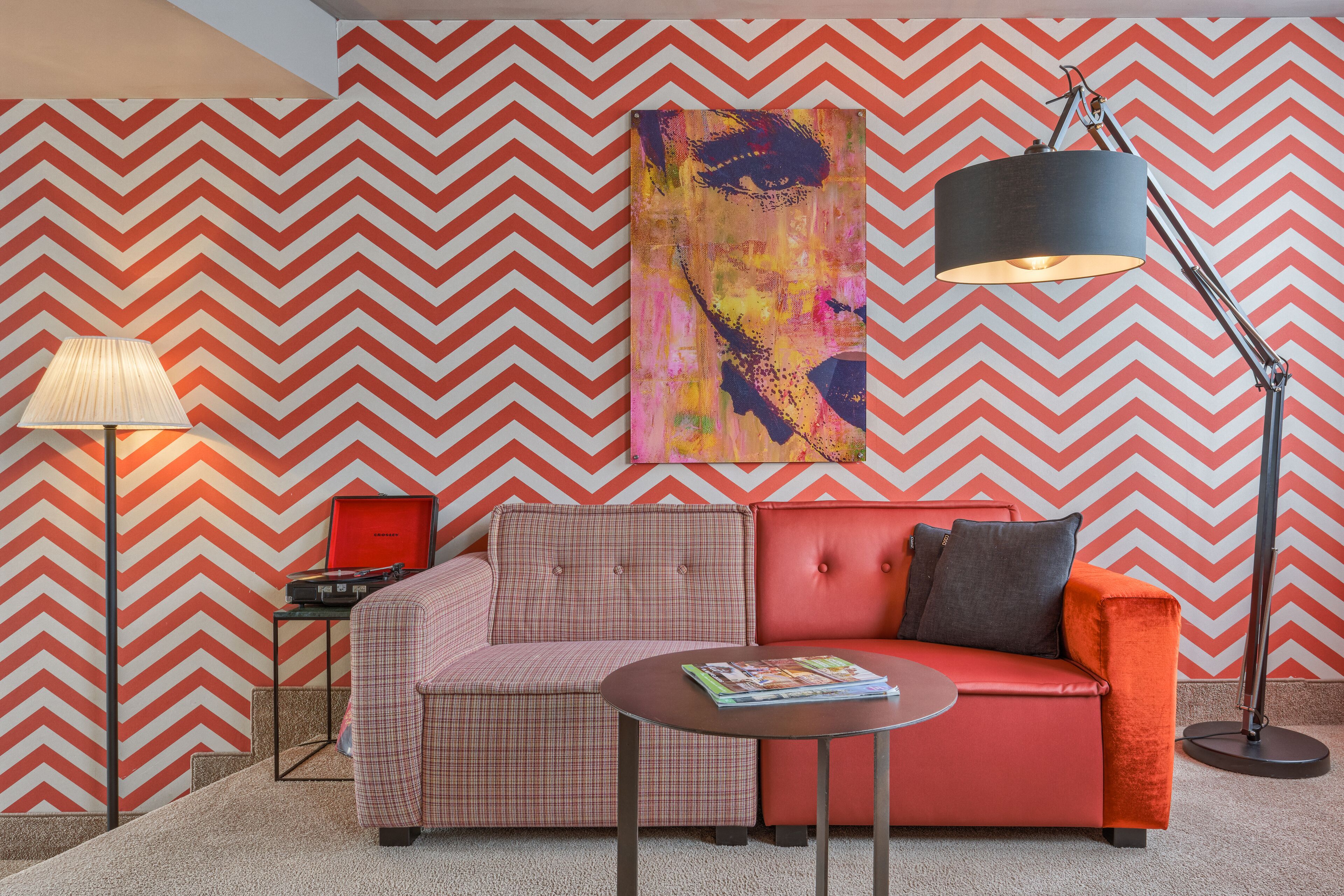 A modern living room with a chevron-patterned wall, a red sofa, a plaid armchair, a floor lamp, and a table with books.