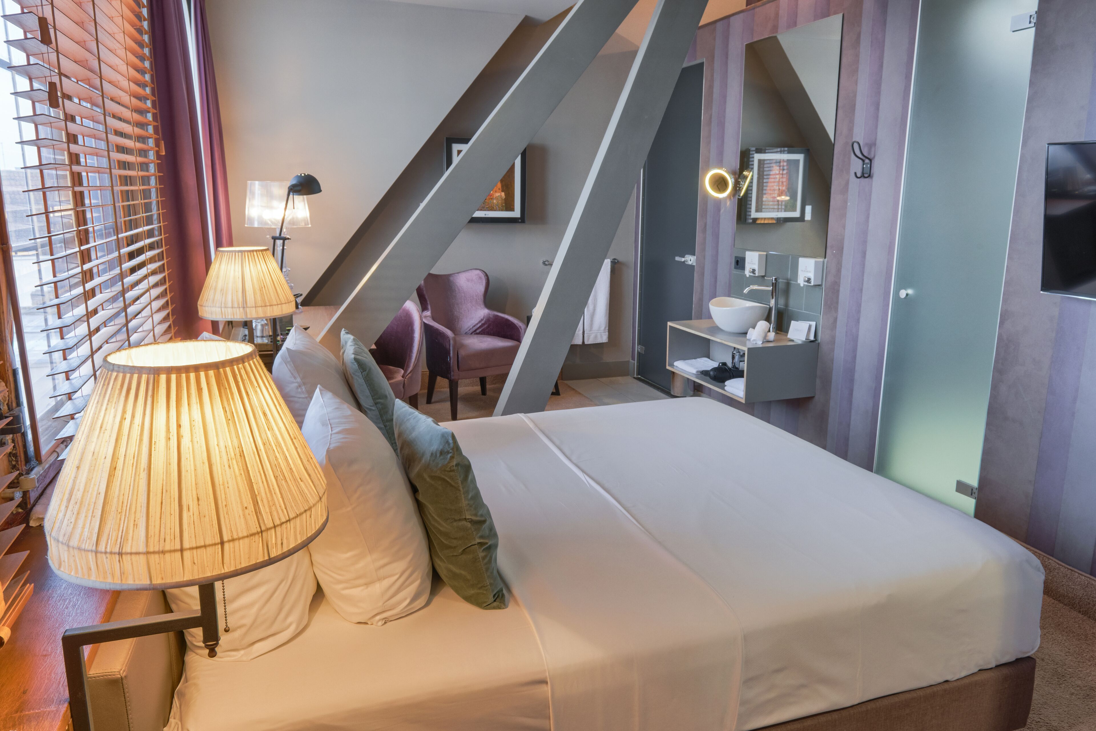 Une chambre moderne avec un lit, deux lampes de chevet, une chaise, une salle de bain avec un lavabo et un miroir, et un escalier.