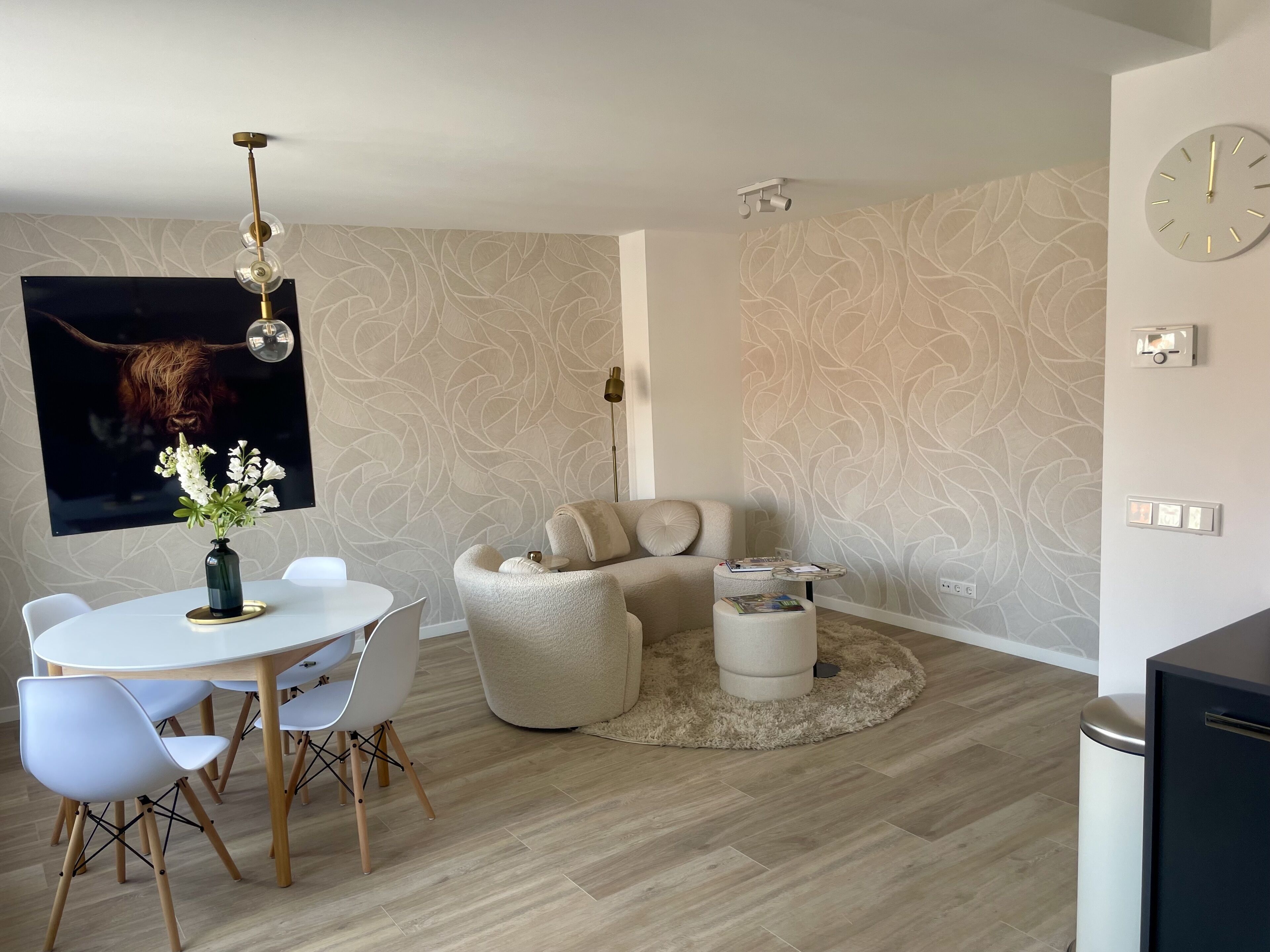 A modern living room with a round dining table, white chairs, a beige sofa, a coffee table, and a mounted clock.
