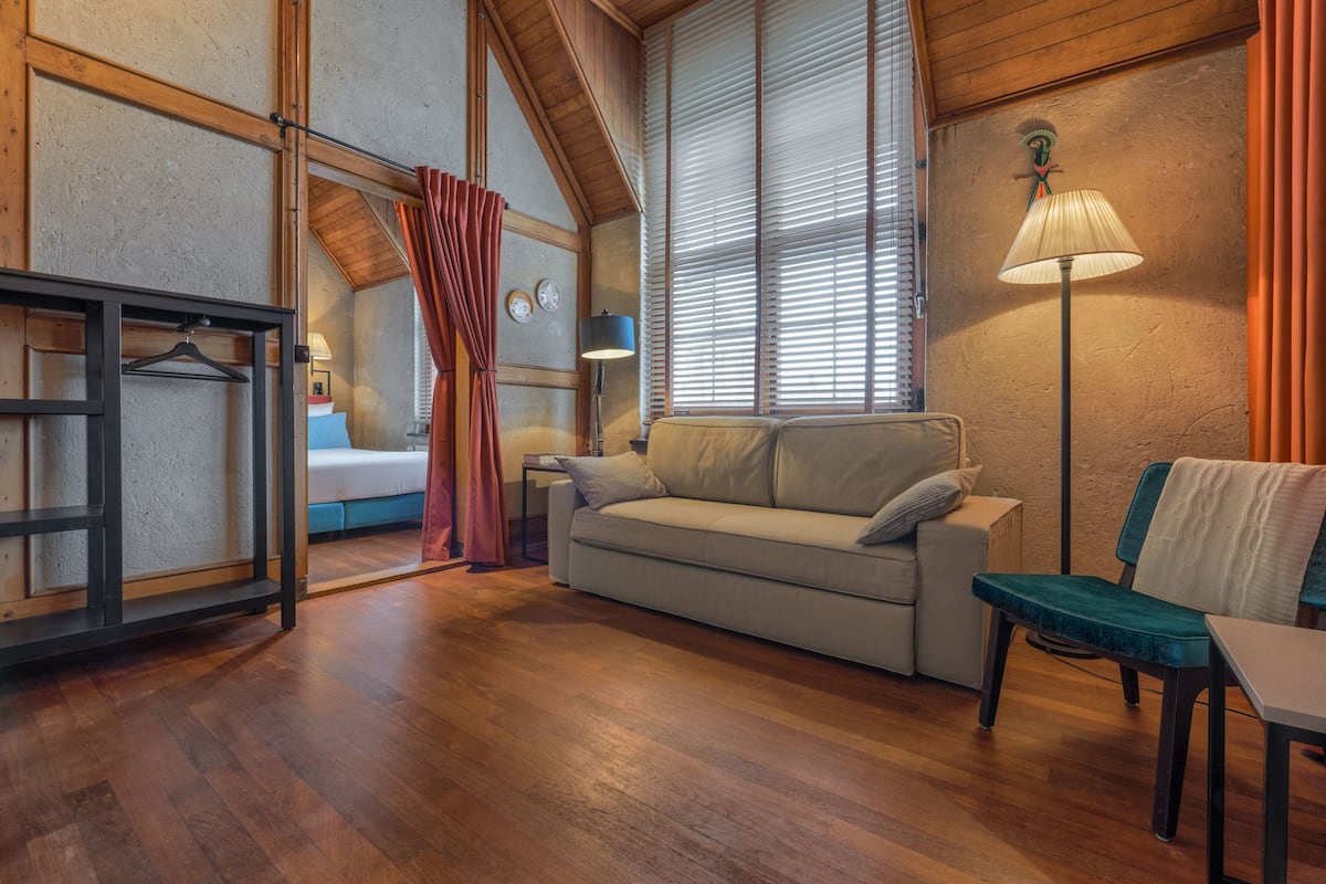 Une pièce confortable avec un canapé, un fauteuil, une table, une lampe et un lit, ainsi qu’une fenêtre et des rideaux.