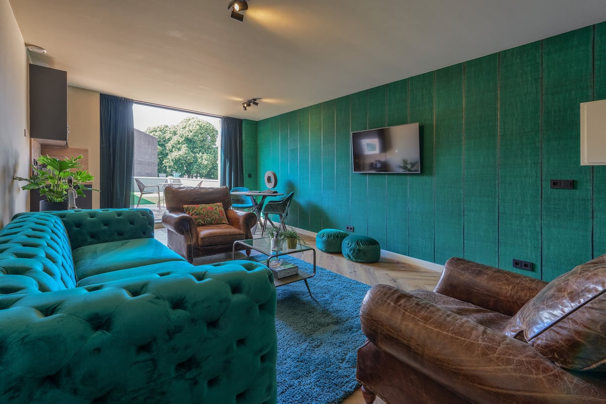 Un salon avec un canapé turquoise, un canapé en cuir marron et une table basse. Un téléviseur est fixé au mur et une plante se trouve sur la gauche.