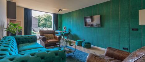 Una sala de estar con un sofá color turquesa, un sofá de cuero marrón y una mesa de centro. Hay un televisor montado en la pared y una planta a la izquierda.