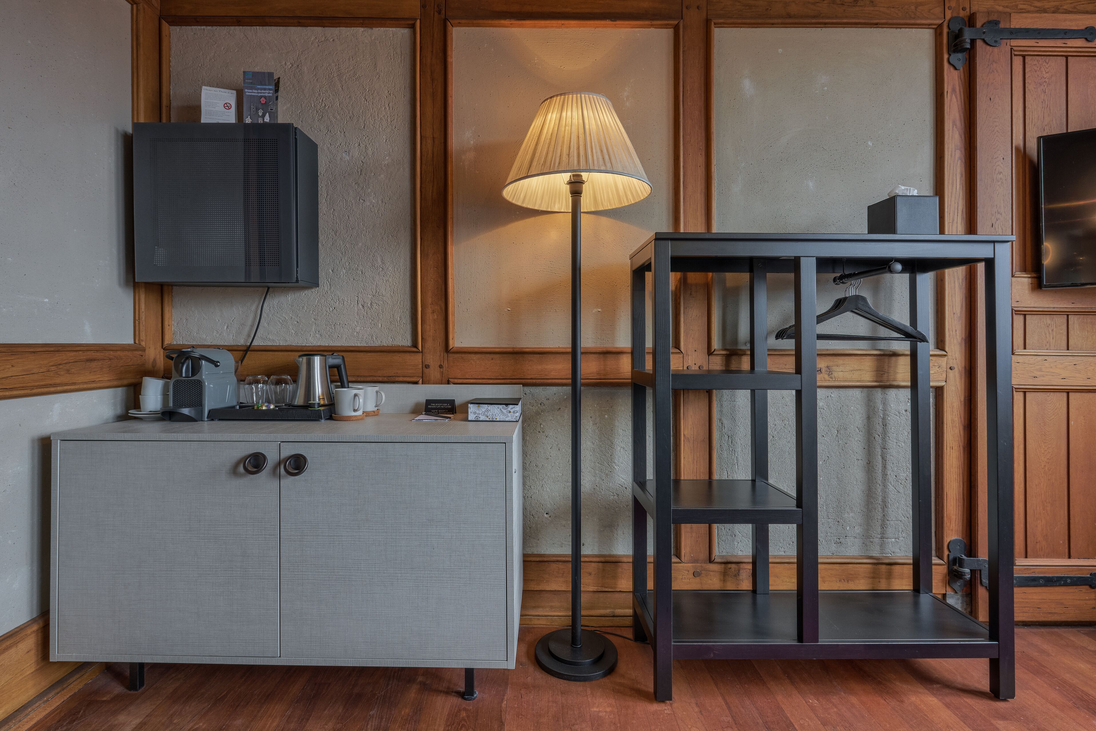Une pièce avec un meuble gris, un lampadaire, un téléviseur fixé au mur et un meuble en bois muni d’un porte-manteau.