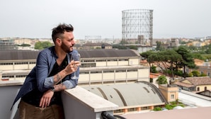 Un hombre apoyado en la barandilla de un balcón, con un paisaje urbano al fondo.