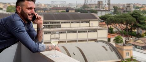 A man leaning on a balcony railing, holding a glass of wine, with a cityscape in the background.