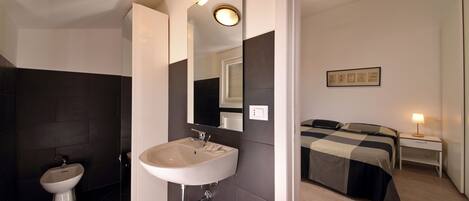A modern bathroom with a sink, bidet, and mirror. There is a bed with a bedside table and lamp in the adjacent room.