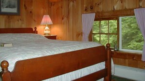 A wooden cabin bedroom with a bed, a bedside lamp, a window with curtains, and a framed picture on the wall. - Fiddler's Green Inn (Stowe)