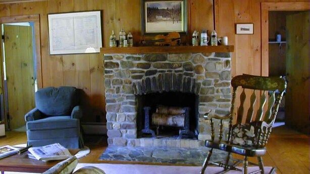 A cozy cabin interior with a stone fireplace, wooden walls, a rocking chair, and a blue armchair. - Fiddler's Green Inn (Stowe)