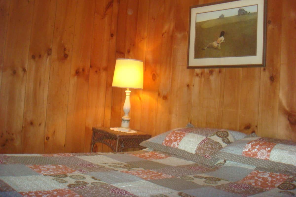 A room with wooden walls, a bed with a floral quilt, a bedside table with a lamp, and a framed picture on the wall.