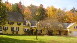 A yellow two-story house with a front porch, a lawn, and trees with autumn foliage in the background. - Fiddler's Green Inn (Stowe)