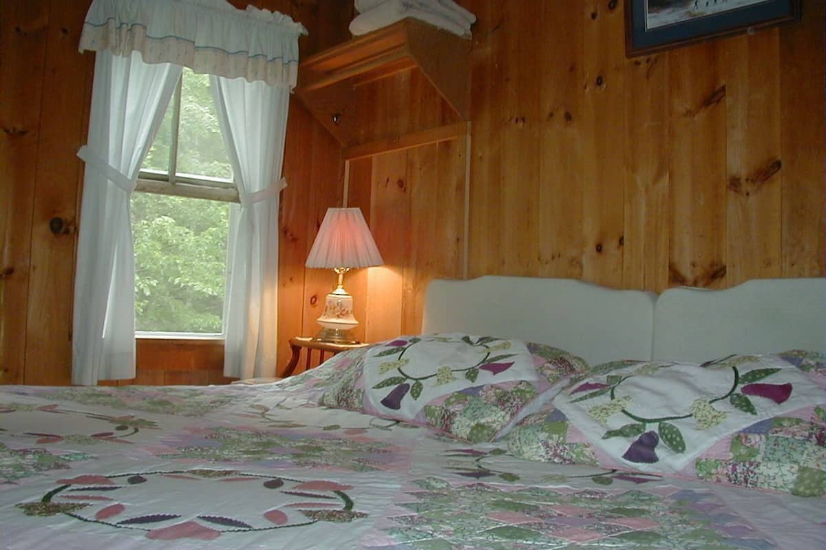 A wooden cabin bedroom with a bed, a nightstand, a lamp, and a window with curtains.