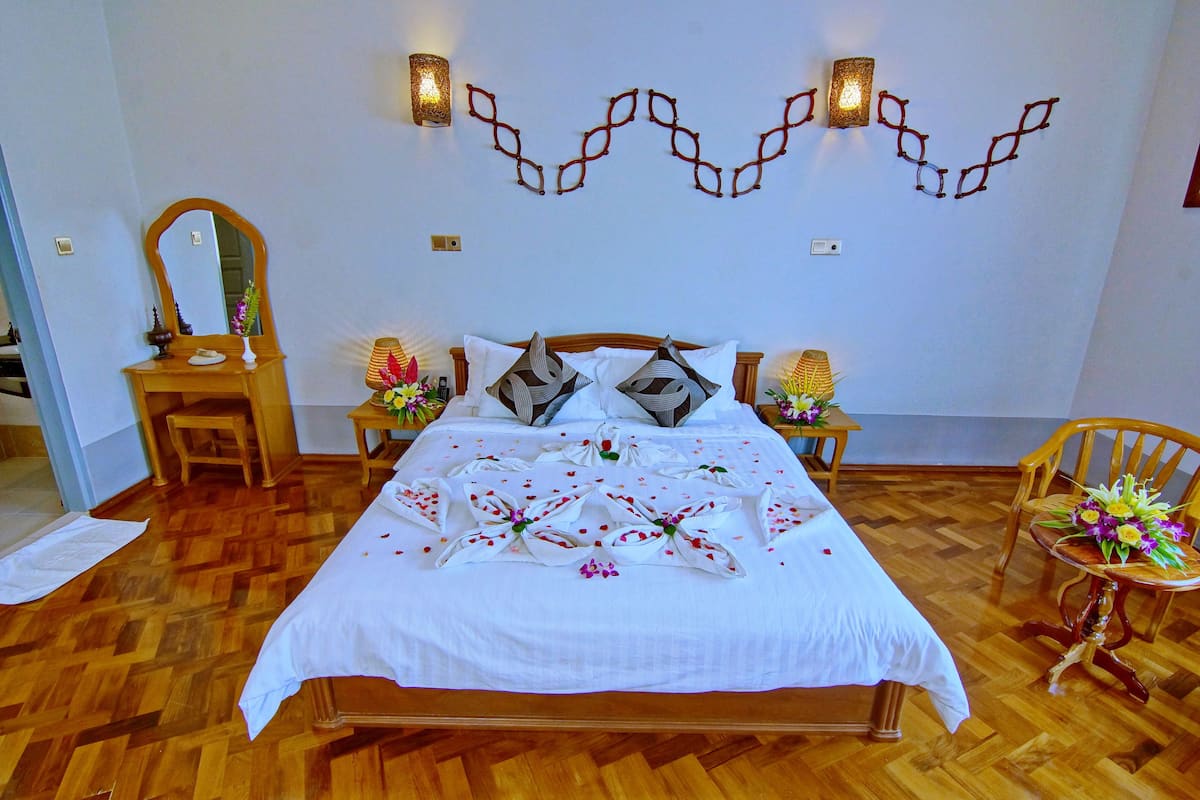 Una cama bien hecha con colcha blanca, arreglos florales decorativos, cabecera de madera y dos mesitas de noche con lámparas y flores.