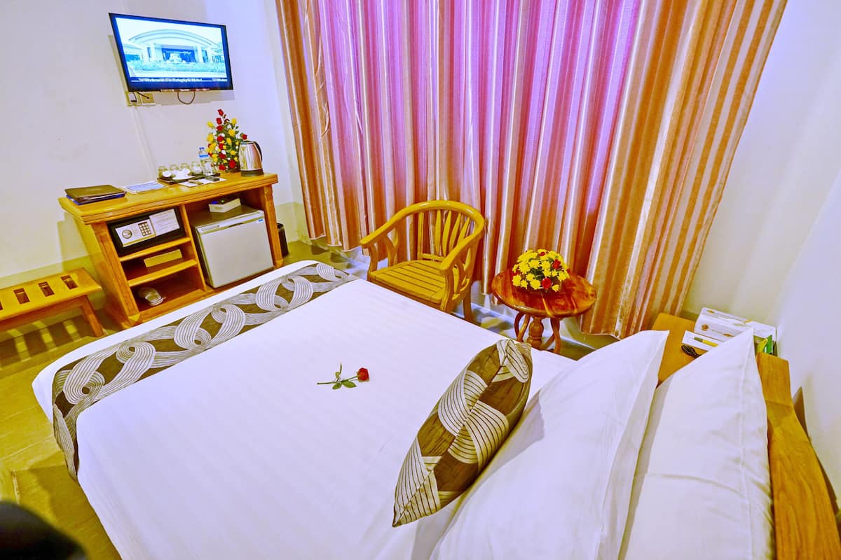 Habitación de hotel con cama, escritorio con computadora, silla, pequeña mesa con un florero y televisor en la pared.