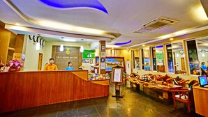 A hotel reception area with a wooden reception desk, a large painting on the wall, and a seating area with wooden chairs and tables. - Triumph Hotel (Mandalay)