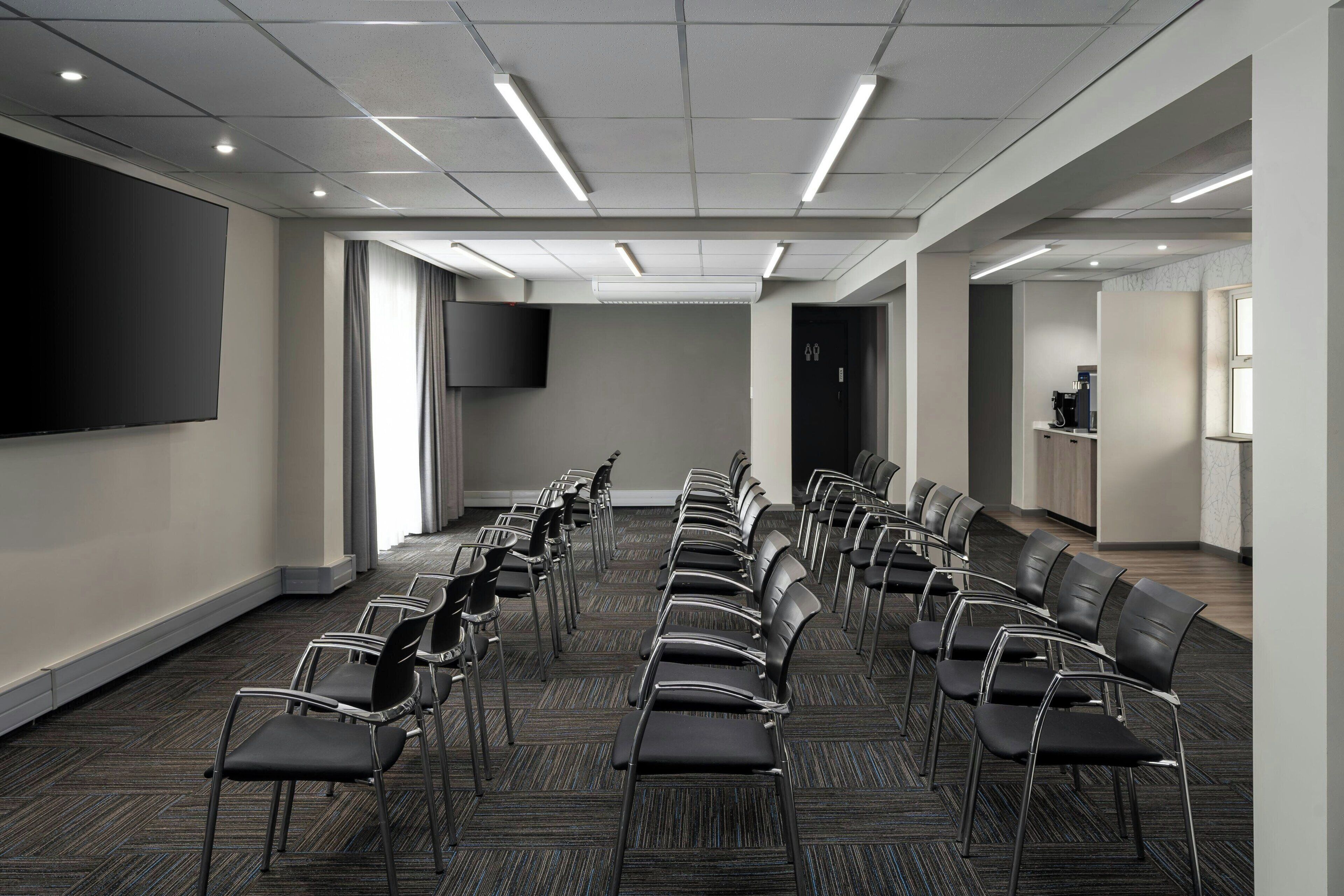 Uma sala de conferências com fileiras de cadeiras, uma tela grande e um espaço para café.