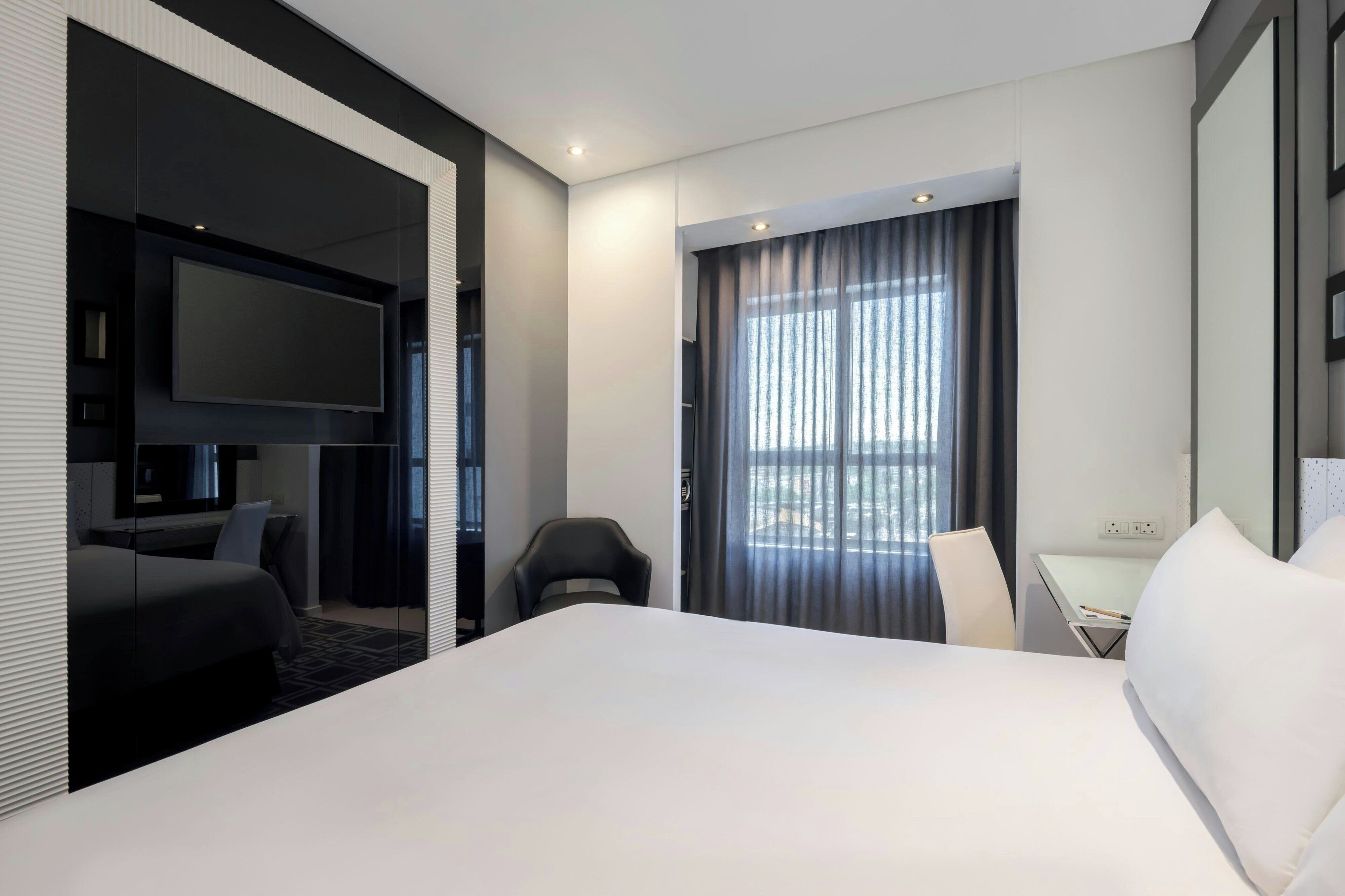 Una habitación de hotel moderna con una cama grande, un escritorio, una silla, un televisor y una ventana con cortinas.