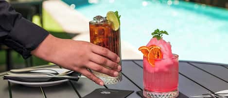 A person holding a glass with a drink, a pink cocktail with an orange garnish, and a pool in the background.