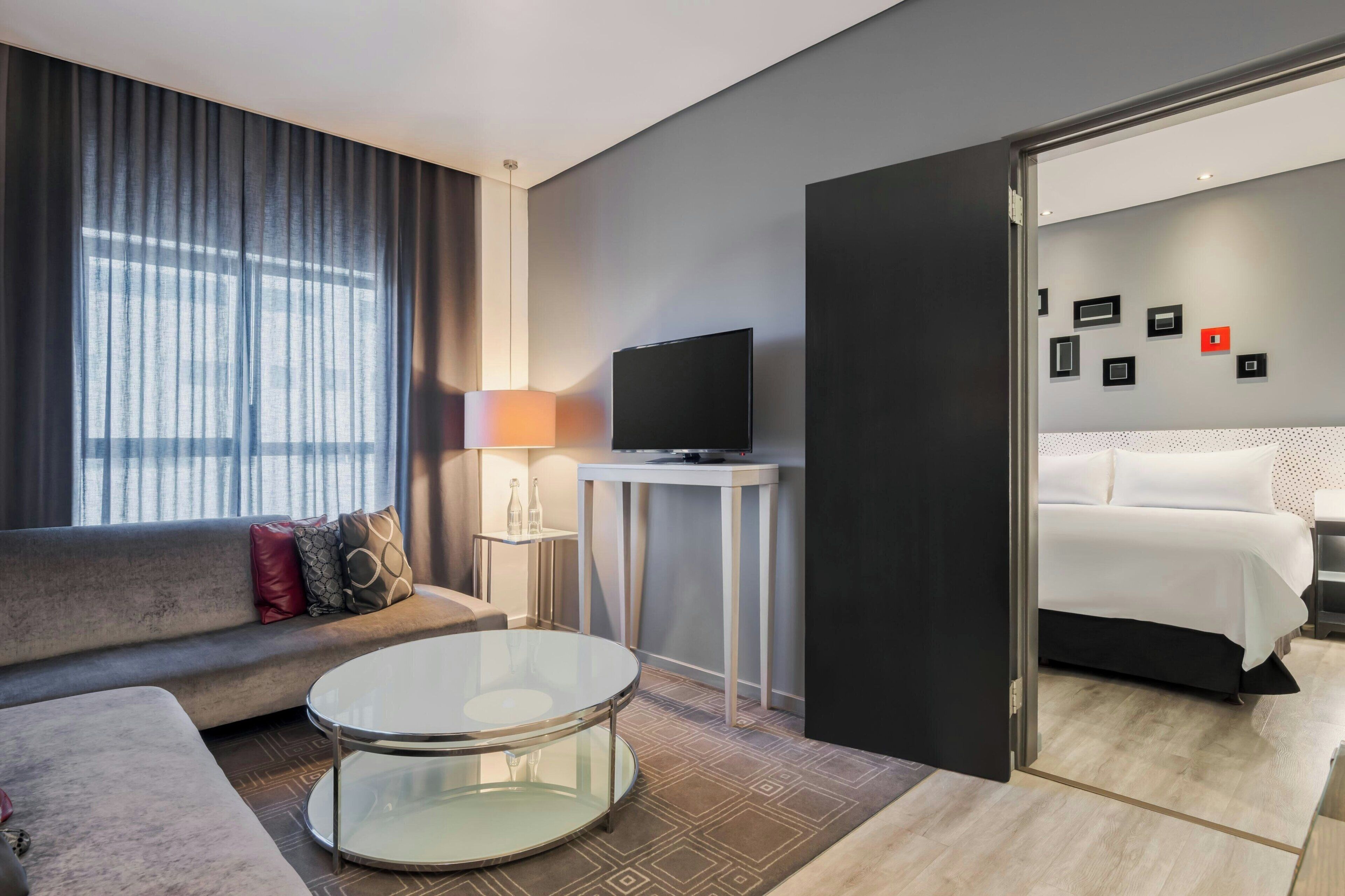 Een moderne hotelkamer met een bank, een rond glazen bijzettafeltje, een televisie, een nachtkastje en een bed.