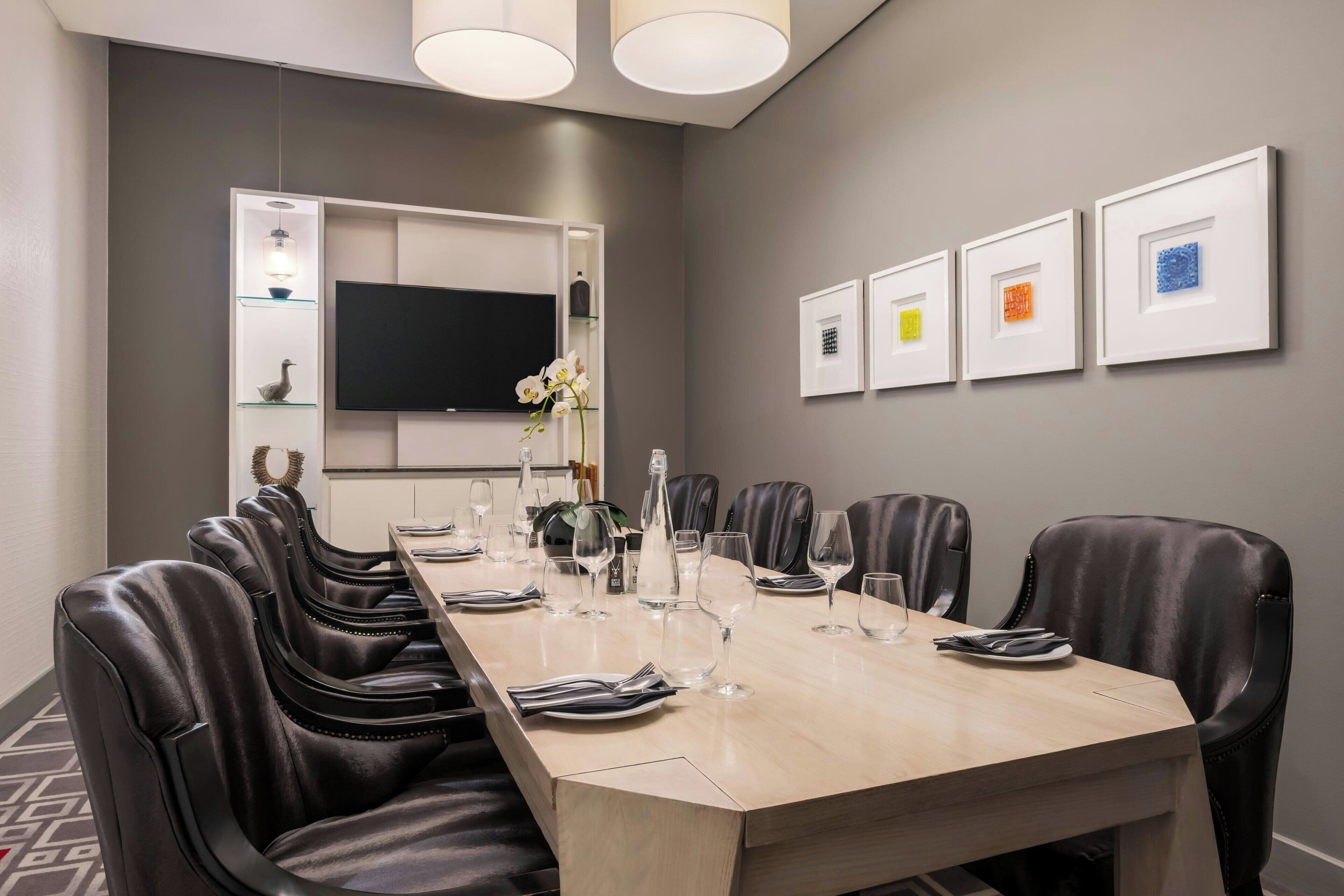Un salón de conferencias con una larga mesa, sillas de cuero, un televisor de pantalla plana, obras de arte enmarcadas y un jarrón con flores.
