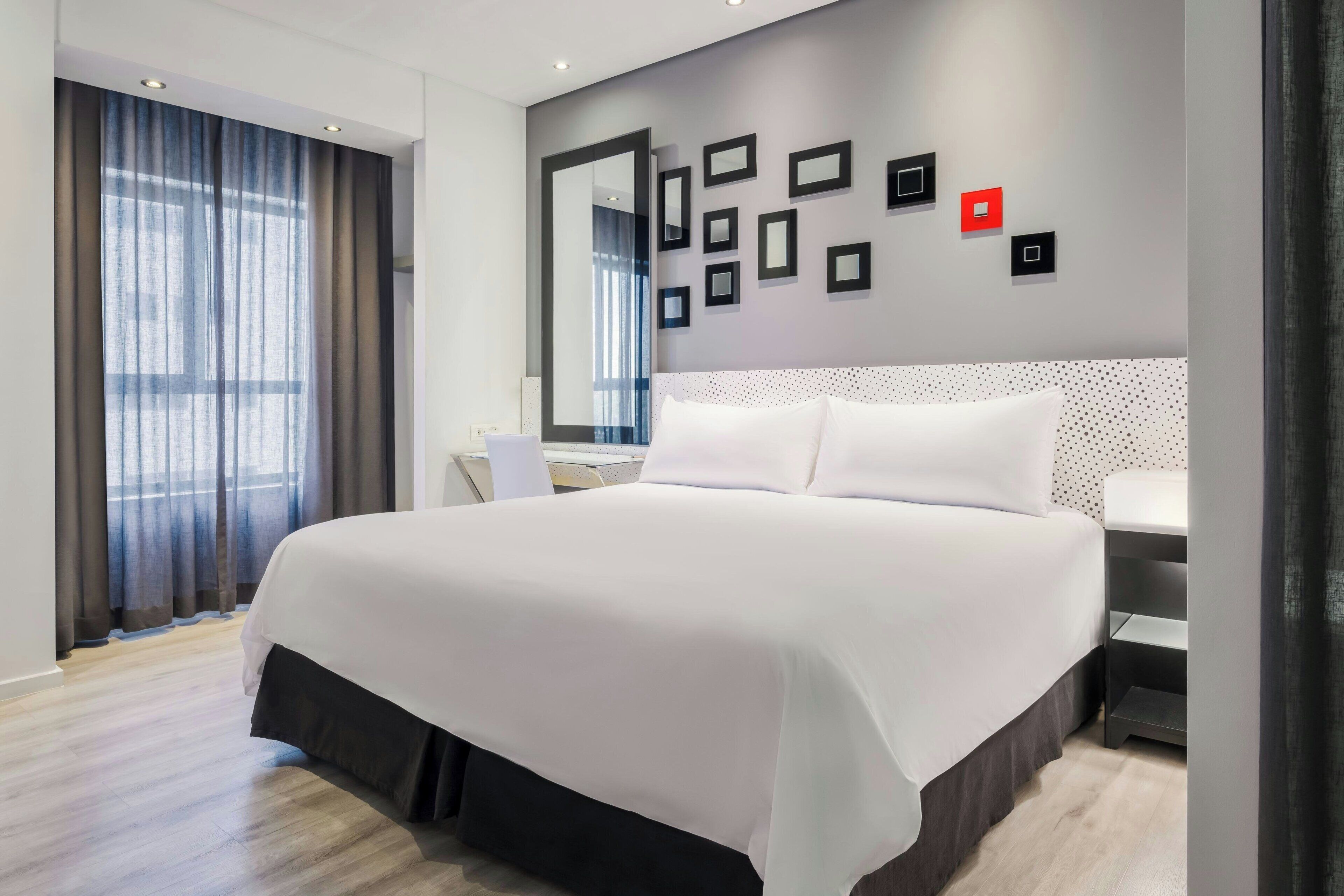 A modern hotel room with a large bed, a desk, a mirror, and a wall with framed pictures.