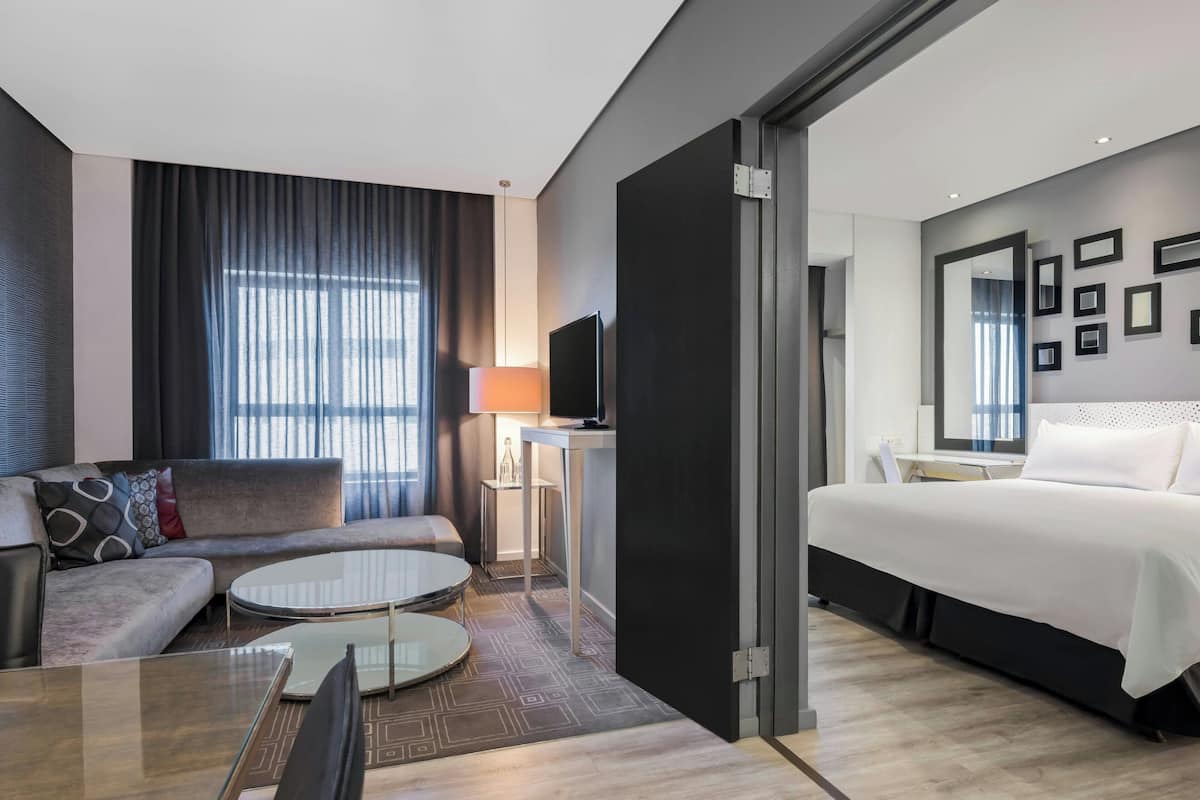 A modern hotel room with a sofa, a round dining table, a bed, a TV, and a desk with a lamp.