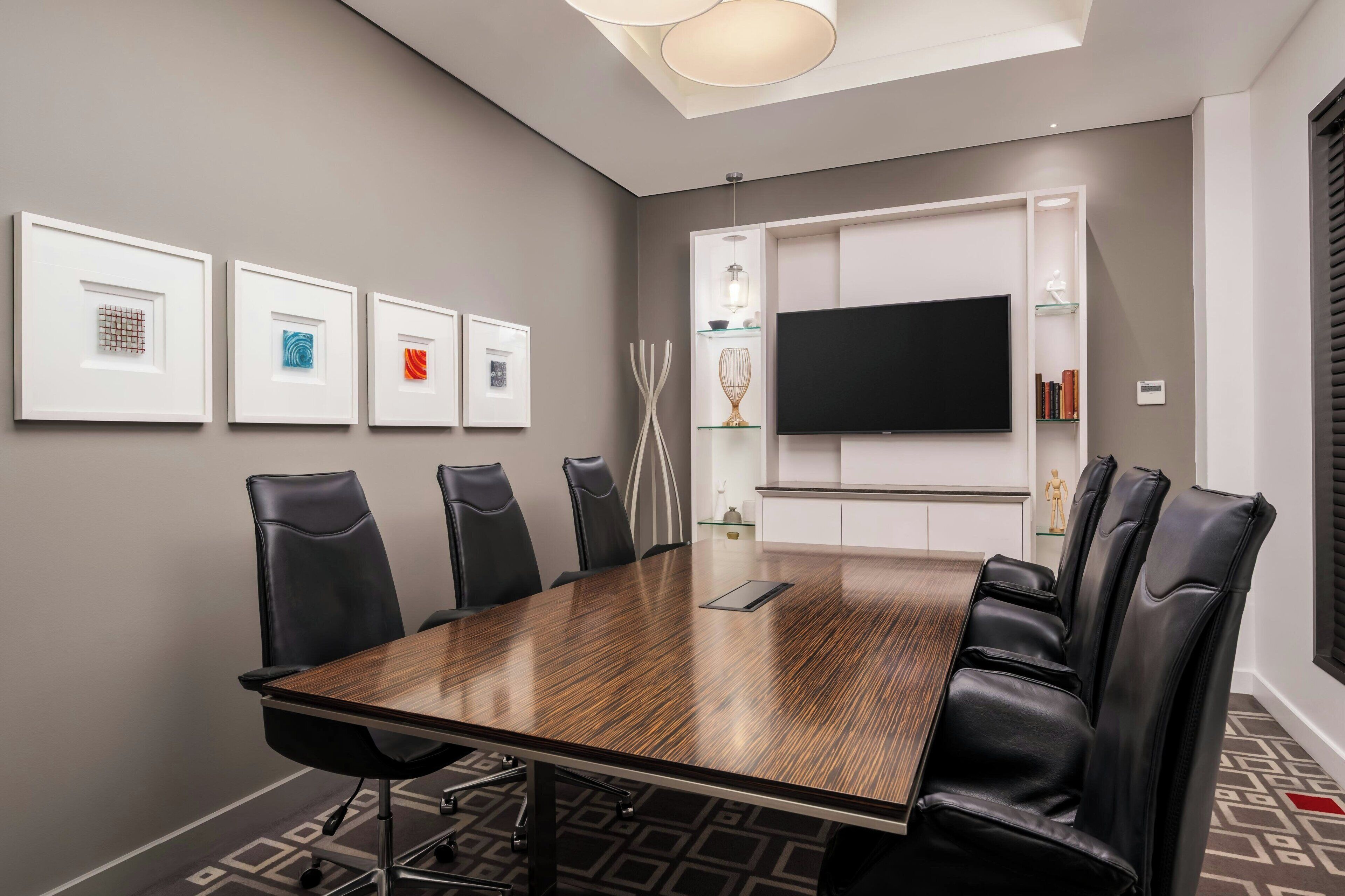 Un salón de conferencias con una larga mesa, sillas de cuero negro, obras de arte enmarcadas en la pared, un televisor de pantalla plana y una lámpara en el techo.