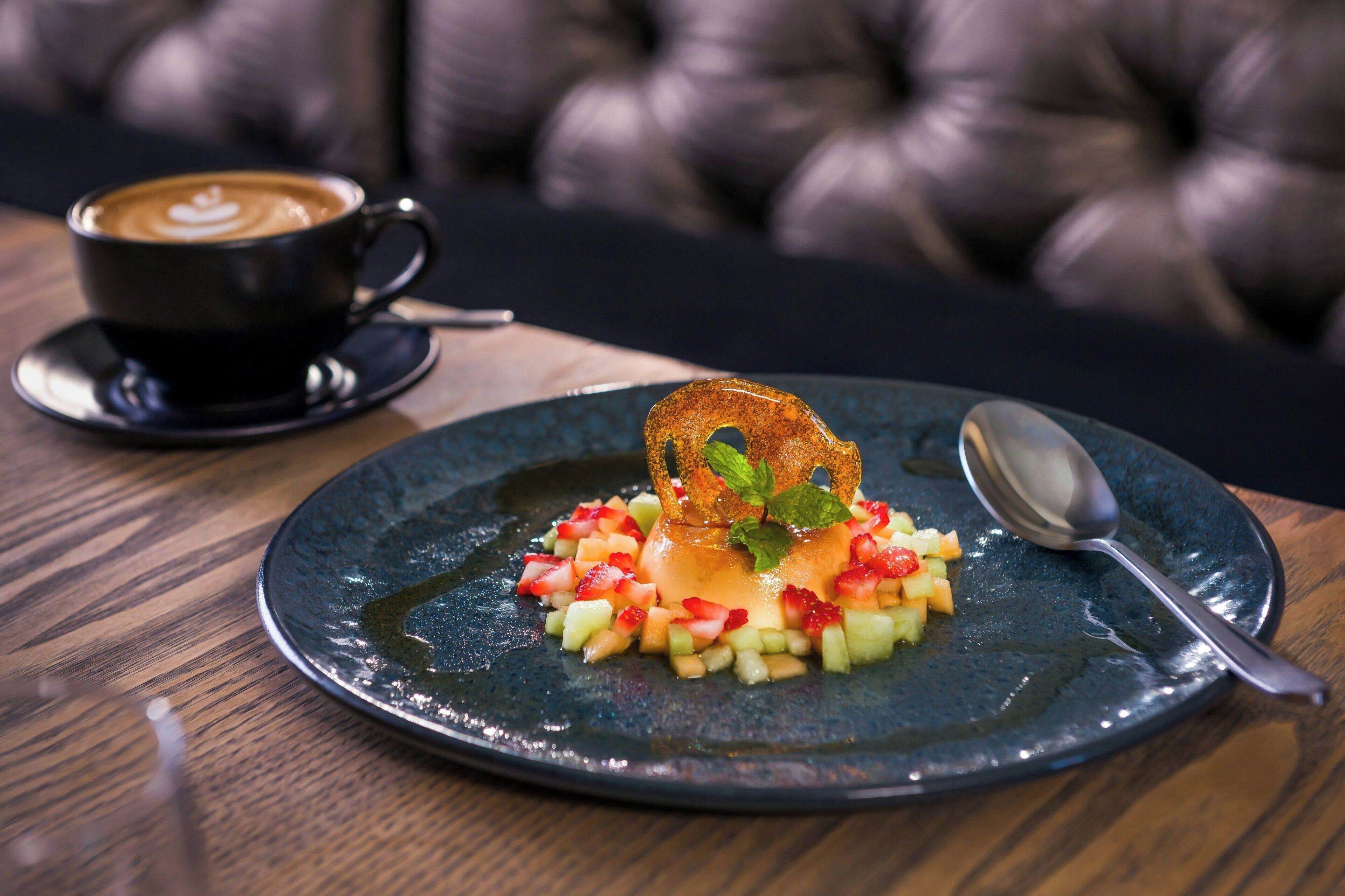 Prato de sobremesa com frutas e um raminho de hortelã sobre um prato de cerâmica escura, acompanhado de uma xícara de café e uma colher em uma mesa de madeira.