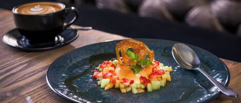 A dessert plate with fruit and a mint garnish on a dark ceramic plate, accompanied by a cup of coffee and a spoon on a wooden table.
