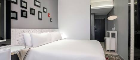 A modern hotel room with a neatly made bed, a wall-mounted picture frame, a bedside table, and a small white nightstand.