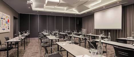 Un salón de conferencias con varias mesas, sillas, una pantalla de proyector y una pizarra grande.