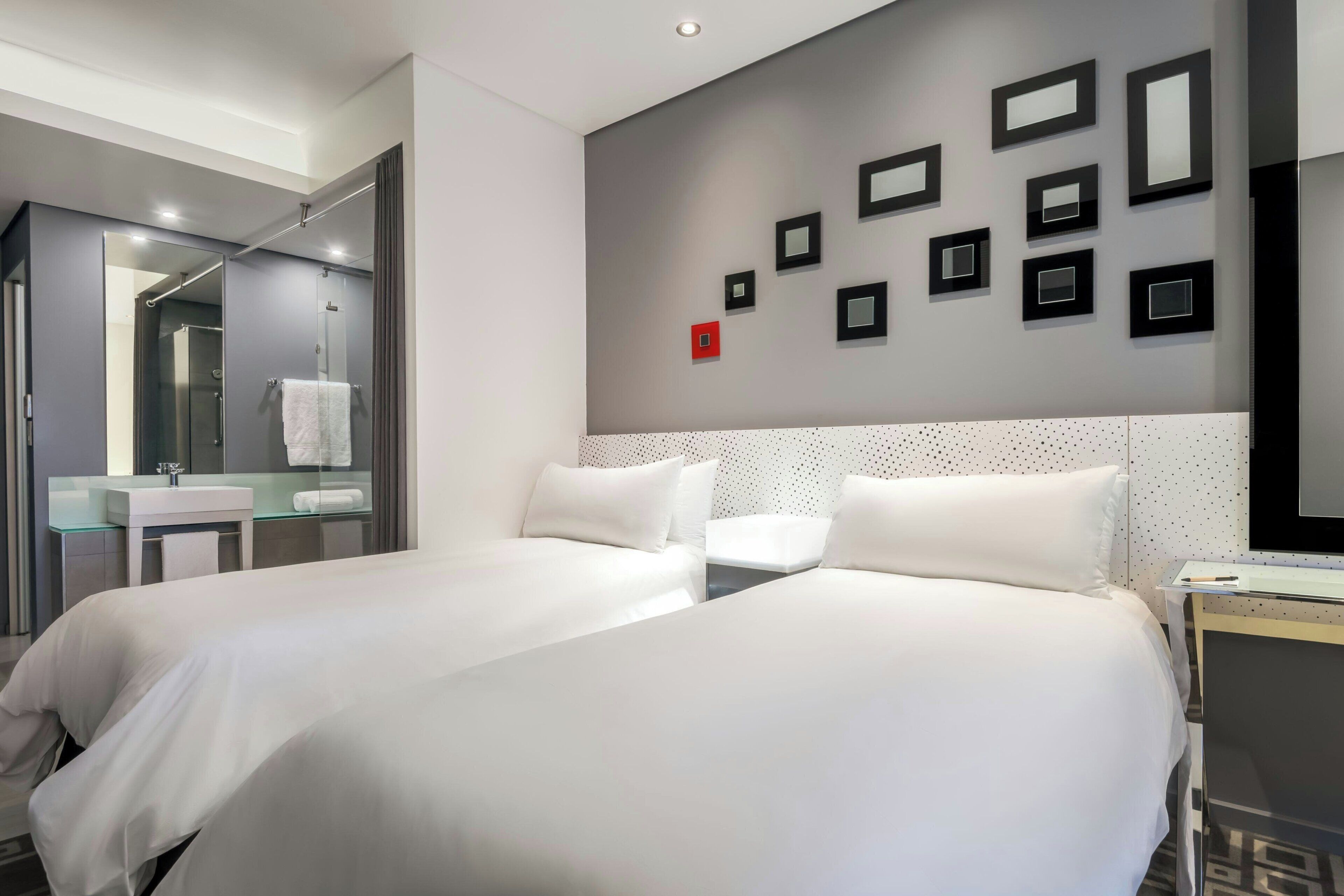 A hotel room with two beds, a glass shower enclosure, a white sink, and a wall with framed pictures.