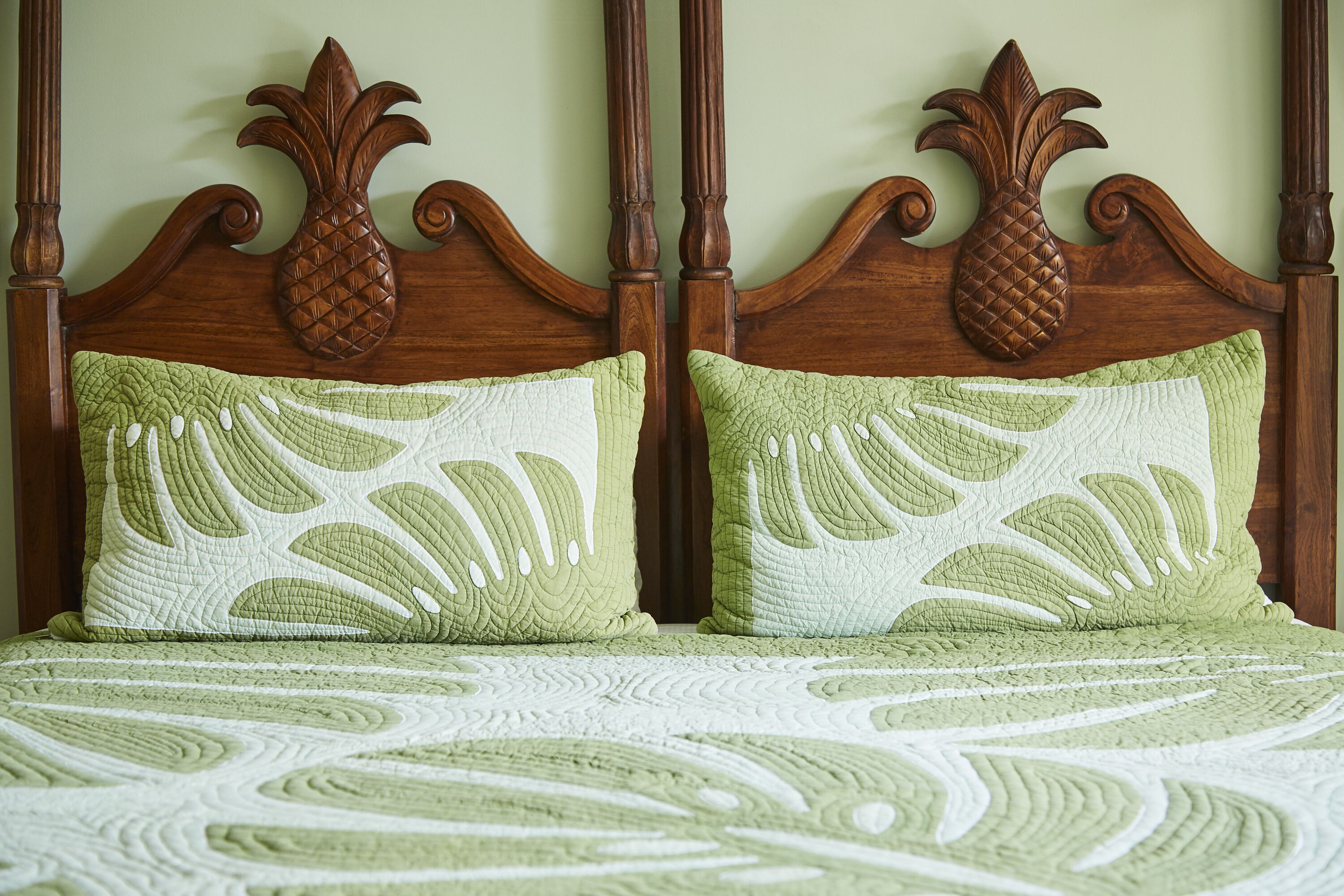 A bed with a wooden headboard and decorative pineapple carvings, two pillows with a leaf pattern, and a green and white quilt.