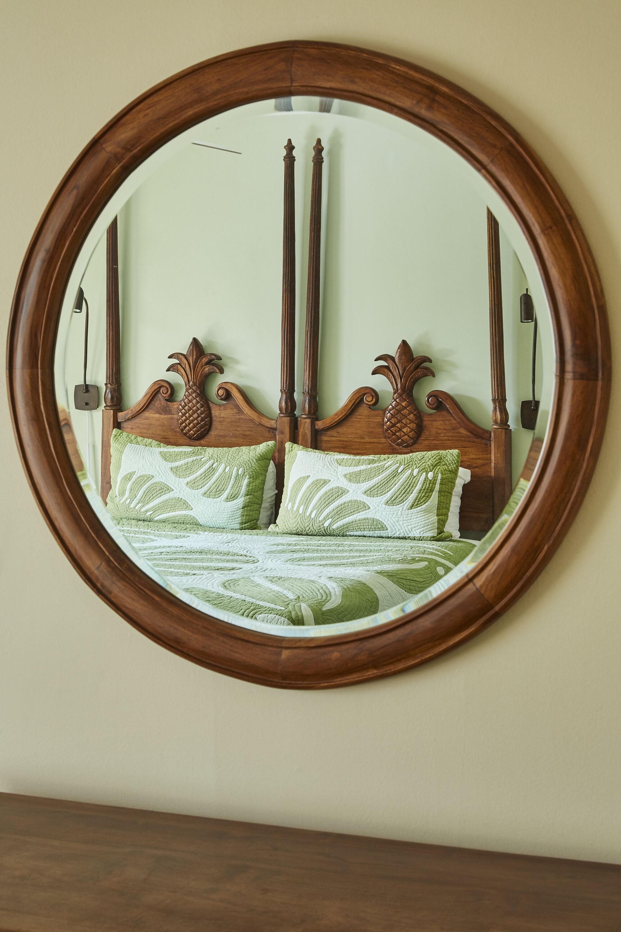Um espelho redondo com moldura de madeira reflete uma cama com cabeceira de abacaxi e roupa de cama com padrão verde.