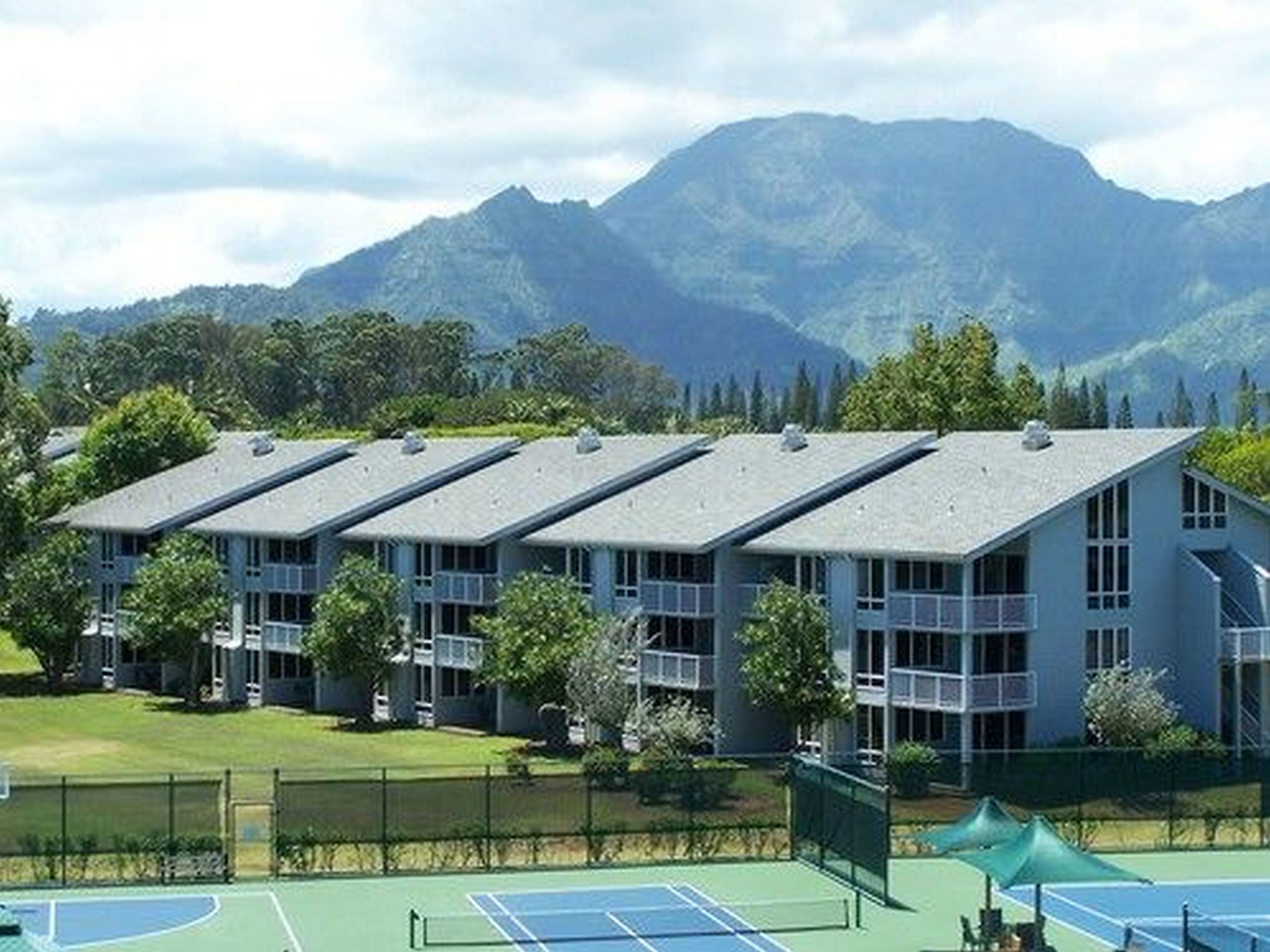 Un hotel de varios pisos, con cancha de tenis y montañas al fondo.