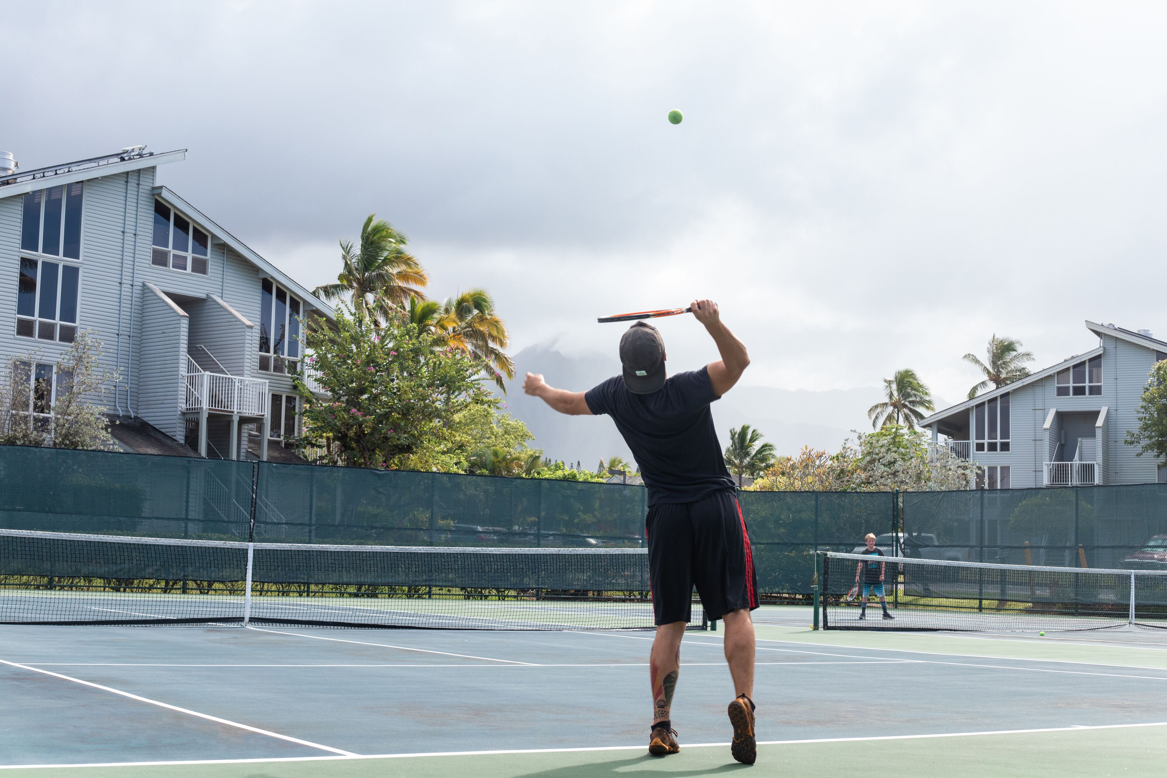 Una cancha de tenis con red, un jugador con camiseta y pantalones cortos negros, y una casa al fondo.
