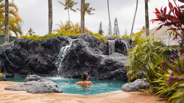 A person in a pool with a waterfall and palm trees in the background. - The Cliffs at Princeville (Princeville)