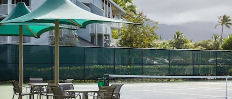 Un court de tennis avec un filet, entouré de chaises et de parasols, et un bâtiment avec des balcons en arrière-plan.