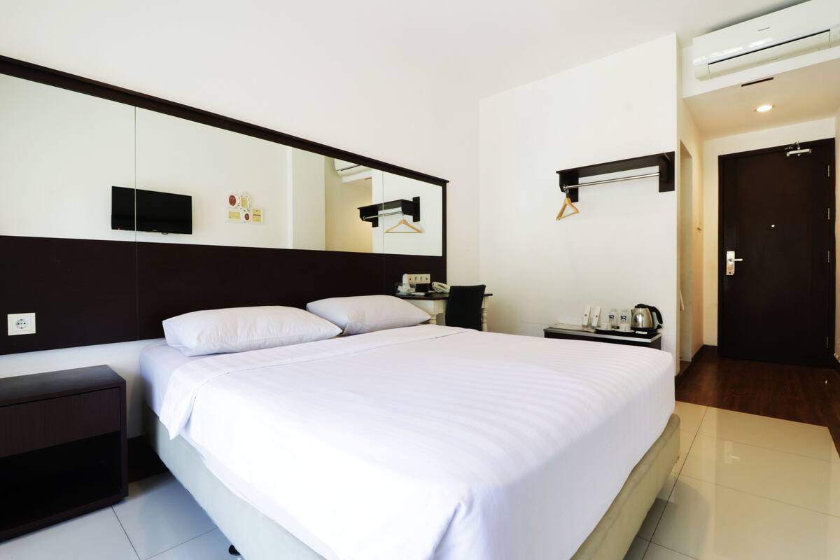 A hotel room with a large bed, a bedside table, a mirror, a TV, a coat rack, and a door.