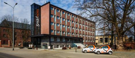 Un hôtel moderne à la façade en briques, doté de nombreuses fenêtres et d’une enseigne bien visible. Deux voitures de la marque SmartHotel sont garées devant l’entrée.
