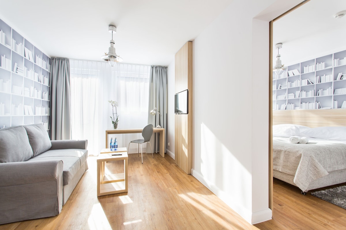 A modern hotel room with a wooden floor, a grey sofa, a desk with a chair, a bookshelf, and a bed with a white duvet.