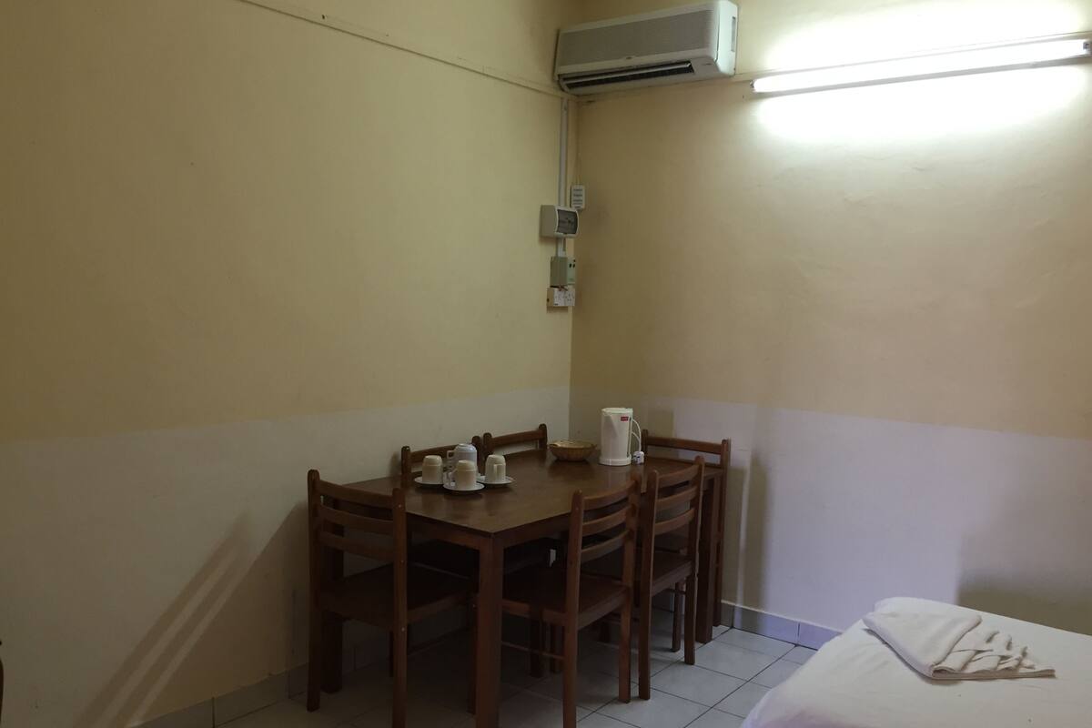 Una habitación pequeña y limpia con una mesa de madera preparada para cuatro personas, una cama individual y un equipo de aire acondicionado en la pared.