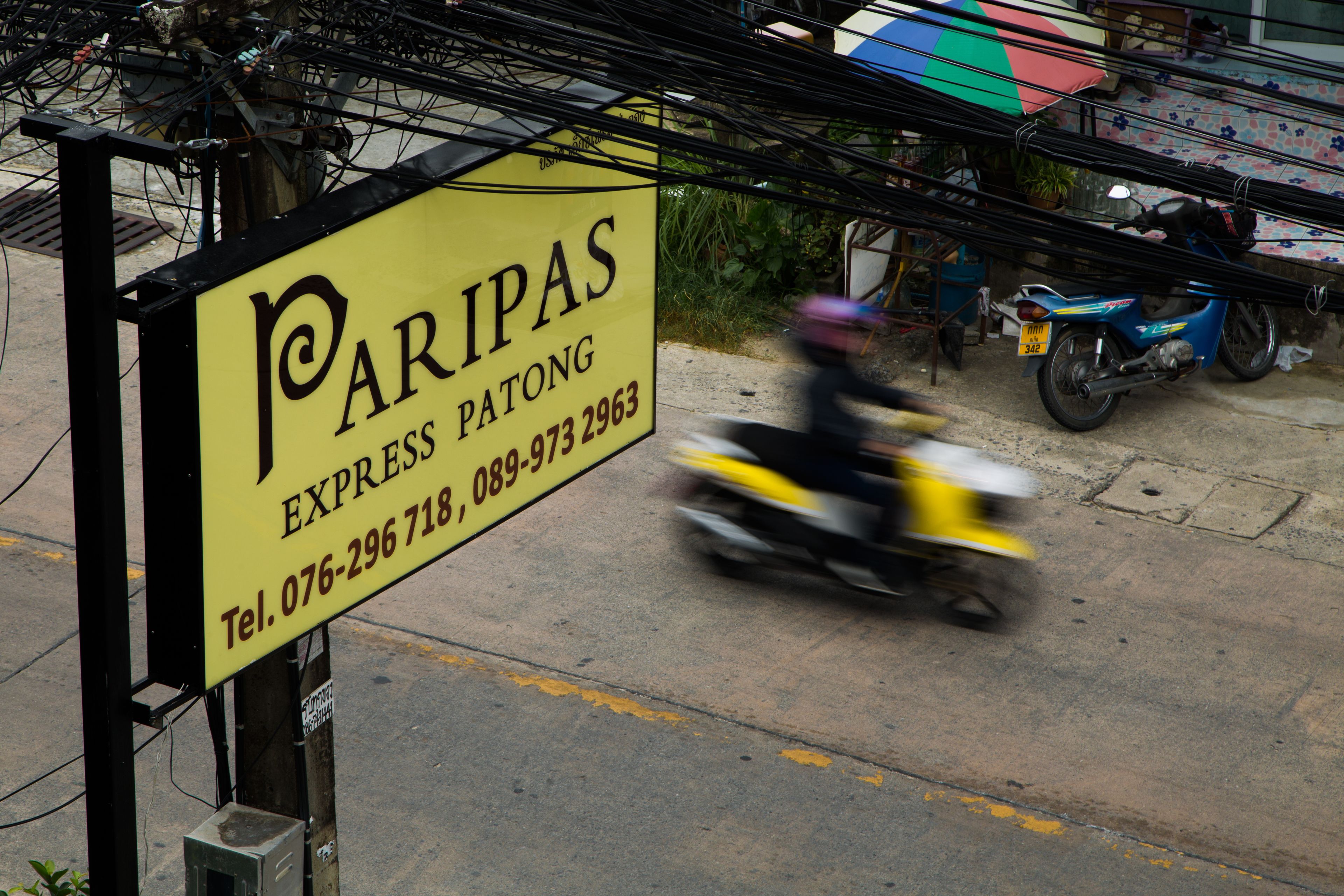 A yellow sign with black lettering for 'Paripas Express Patong' and a contact number.