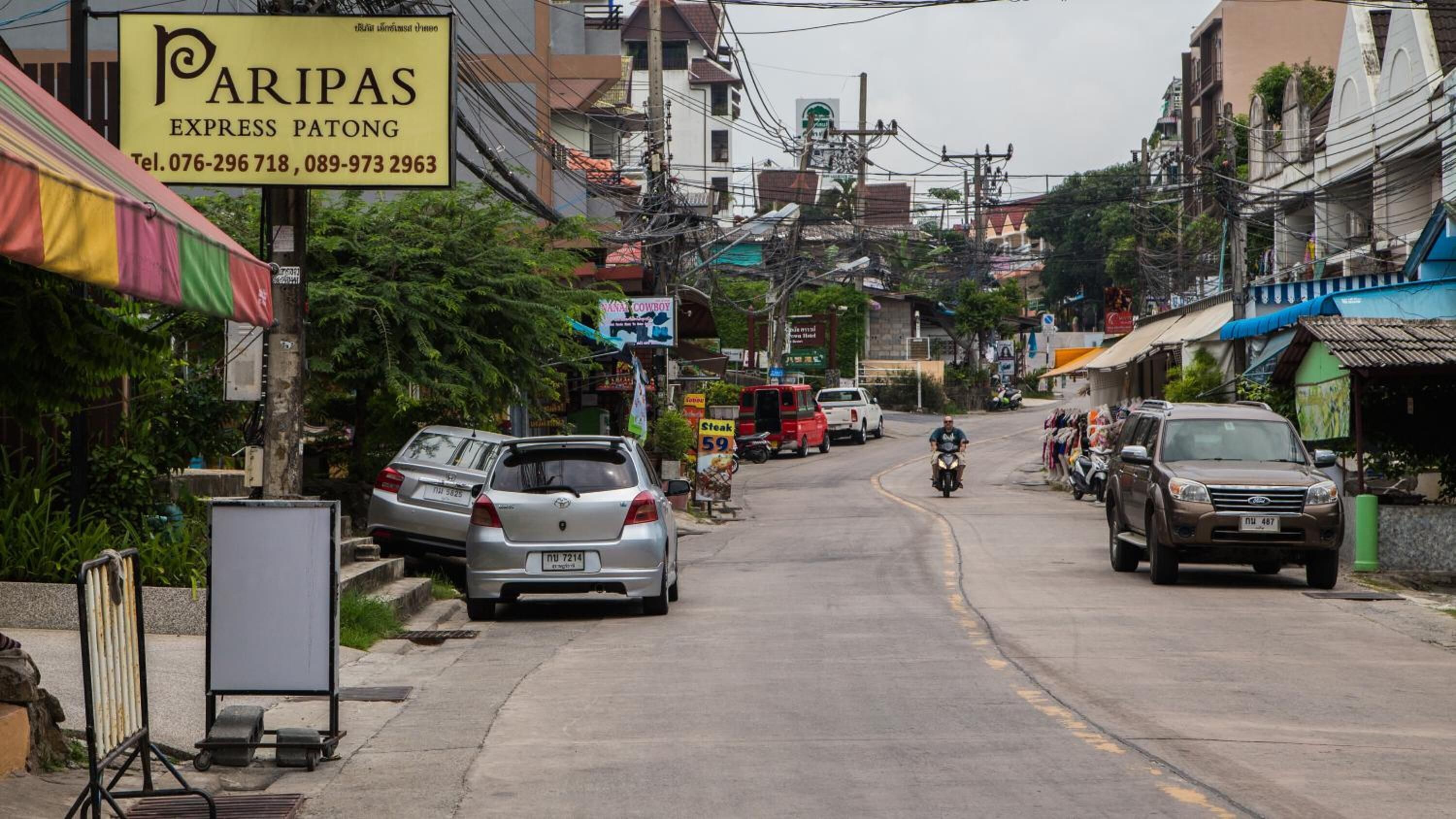 Vista de la calle con coches estacionados, una motocicleta y un cartel que dice 'Paripas Express Patong' con números de contacto.