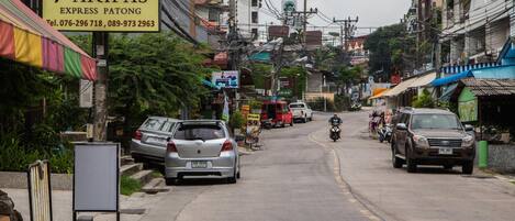 Vista de la calle con coches estacionados, una motocicleta y un cartel que dice 'Paripas Express Patong' con números de contacto.