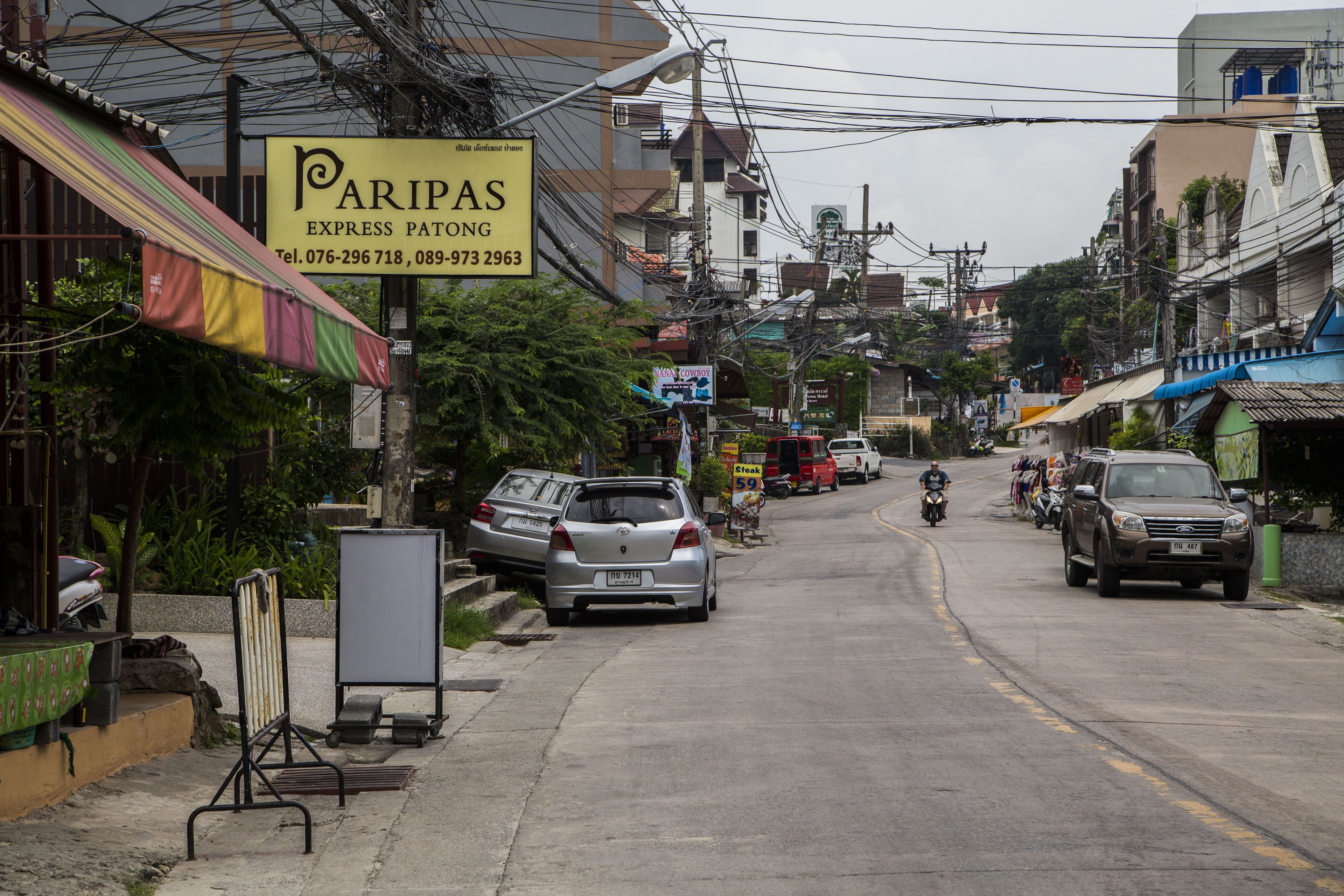 A street with parked cars and motorcycles, a sign for 'Paripas Express Parking', and buildings with visible power lines.