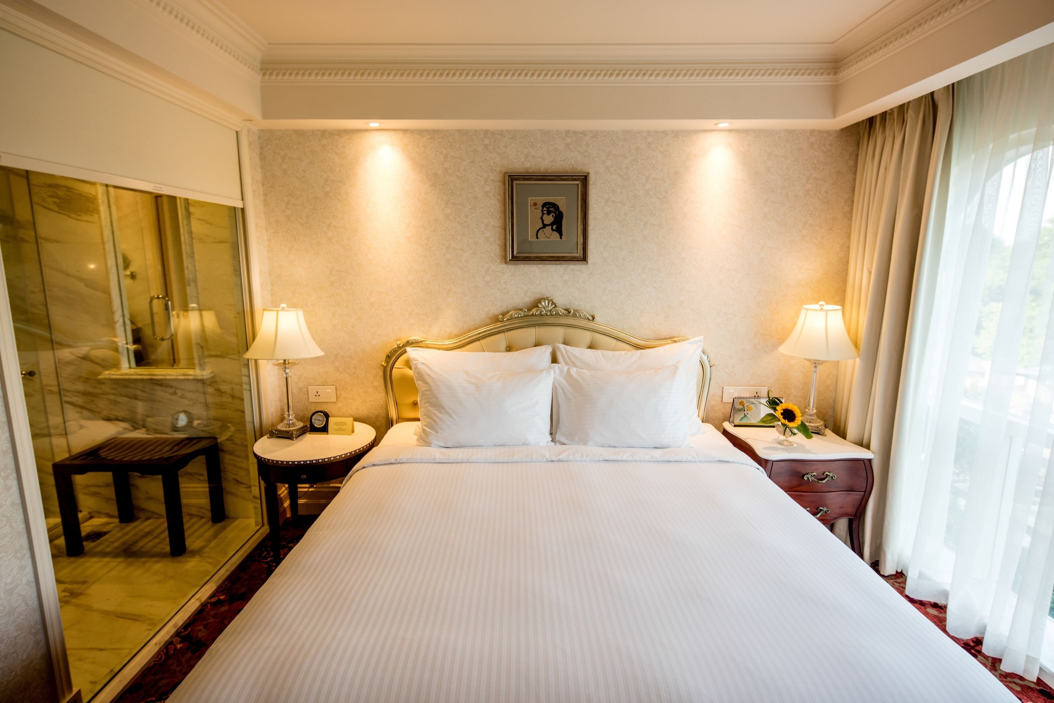 Quarto de hotel com uma cama grande, duas mesas de cabeceira, um criado-mudo, um espelho e uma janela com cortinas.