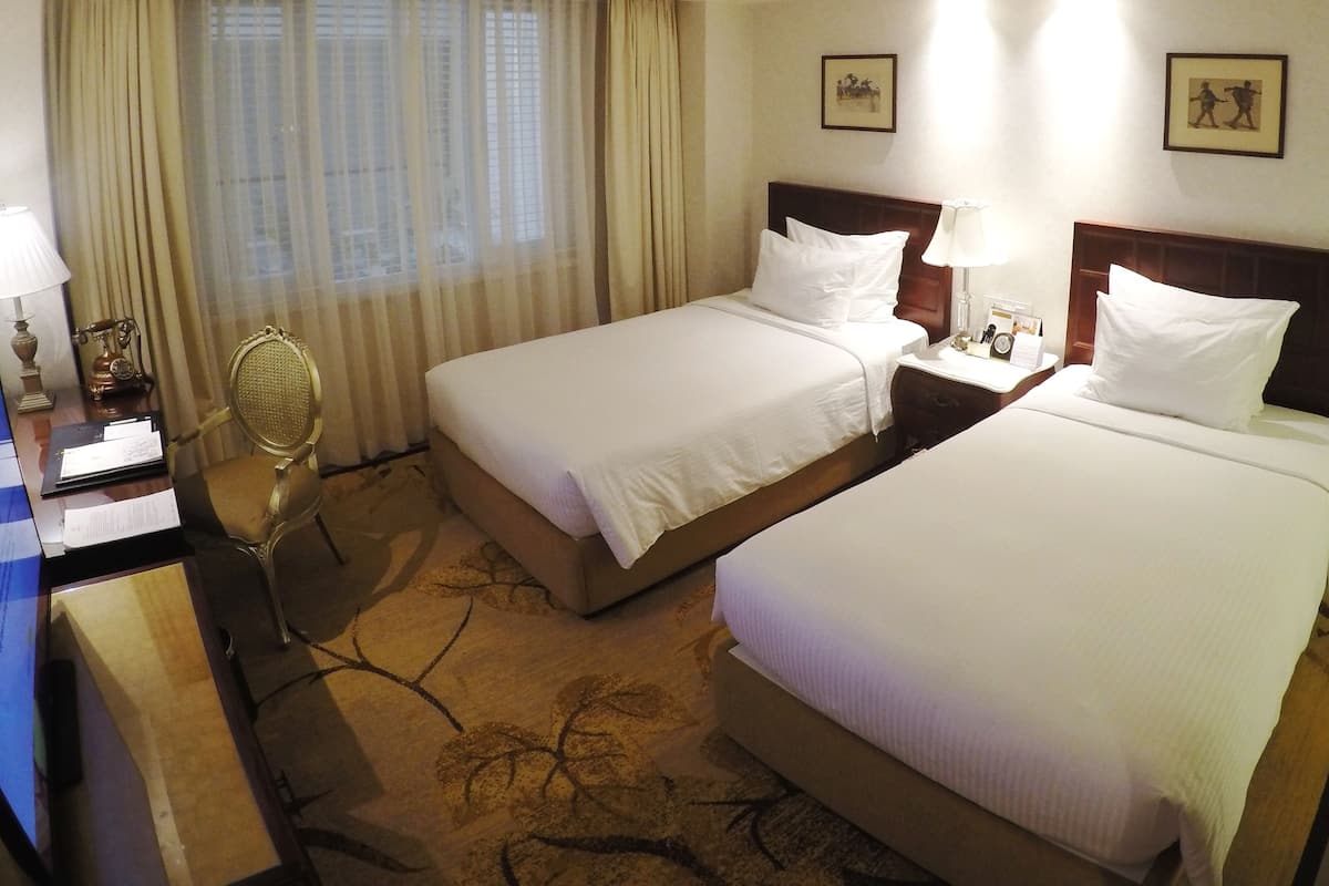 A hotel room with two beds, a desk with a computer, a chair, a desk lamp, and a small table with a lamp and a clock.